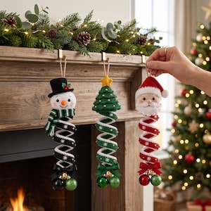 May include: Three hanging crochet ornaments featuring a snowman in a black hat, a green Christmas tree with a yellow star, and a Santa Claus figure. Each ornament has a spiral body with small beads, a satin bow, and two small jingle bells at the base.