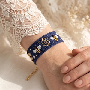 May include: A dark blue woven bracelet featuring a repeating pattern of yellow and black striped bees and gold-colored honeycomb shapes. The bracelet includes a gold-toned metal chain extender with a small round bead at the end of the clasp.