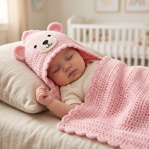 Puede incluir: Una manta con capucha tejida a ganchillo en color rosa claro que presenta un diseño de cara de oso en la capucha con orejas pequeñas y redondeadas, ojos bordados en negro y una nariz. La manta tiene un borde festoneado de hilo suave.