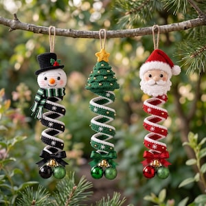 May include: Three hanging crochet ornaments featuring a snowman in a black hat, a green Christmas tree with a yellow star, and Santa Claus. Each figure has a long, spiraling body decorated with white star patterns, a bow, and two small jingle bells.