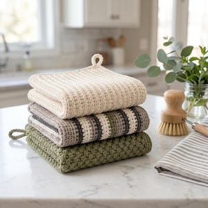 May include: A stack of four crocheted kitchen washcloths in cream, gray, and green. The top washcloth is cream-colored with a loop for hanging. The middle washcloths have striped patterns. The bottom washcloth is green.