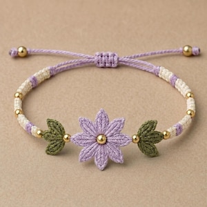 May include: A woven bracelet featuring a central lavender flower with a gold bead center, flanked by two green leaf-shaped accents. The band consists of alternating cream and lavender cord segments interspersed with small gold beads and a sliding knot.