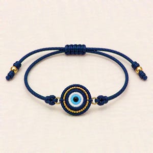 Könnte beinhalten: Ein dunkelblaues Kordelarmband mit einem zentralen, runden Schutzaugen-Anhänger. Das Motiv besteht aus konzentrischen Ringen in Schwarz, Weiß und Hellblau, umrandet von kleinen goldfarbenen Perlen. Das Armband hat einen verschiebbaren Knotenverschluss.
