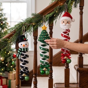 May include: Three crocheted Christmas ornaments hanging from a wooden banister. One is a snowman with a black hat, one is a green Christmas tree with a yellow star, and one is a Santa Claus. Each features a spiral body with small ornaments.