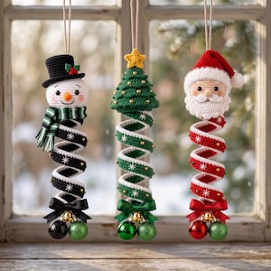May include: Three hanging crochet ornaments featuring a snowman in a black hat, a green Christmas tree with a yellow star, and a Santa Claus head. Each figure has a long, spiraling body decorated with white snowflake patterns and small beads, finished with a satin bow and two small round ornaments at the bottom.
