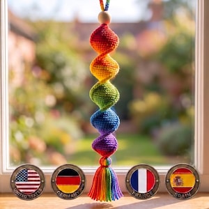 Puede incluir: Un adorno de ganchillo colgante con una espiral vertical de cinco esferas en rojo, naranja, amarillo, verde, azul y morado, rematado con una cuenta de madera y una borla multicolor, situado sobre cuatro fichas metálicas con banderas.