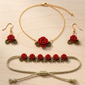 Könnte beinhalten: Ein Schmuckset bestehend aus einer goldfarbenen Kette mit einer zentralen roten Häkelrose und grünen Blättern, passenden Ohrhängern mit roten Rosenmotiven und einem beigen Kordelarmband mit sechs kleinen roten Häkelrosen.