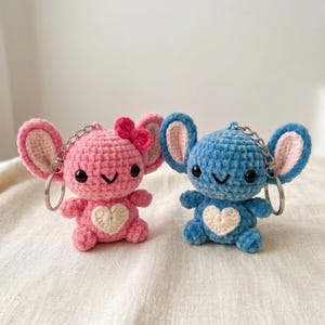 May include: Two crocheted mouse keychains. One is pink with a pink bow and the other is blue. Both have large ears, black eyes, and a white heart on their bellies. Each keychain has a silver chain and ring.