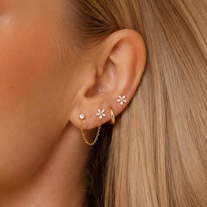 May include: A set of gold-toned earrings featuring a small flower stud, a single round clear stone stud, and a thin gold chain connecting the two. A small gold hoop earring and a tiny flower stud are also worn in the upper ear cartilage.