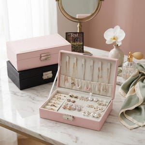 May include: A pink jewelry box is open, displaying necklaces, rings, and earrings. A closed pink box and a black box are stacked behind it. The jewelry box is on a marble surface, with a mirror and a perfume bottle in the background.
