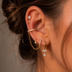 Puede incluir: Un conjunto de pendientes y puños dorados. El conjunto incluye un pendiente en forma de flor con una lágrima colgante, un puño de oreja conectado por una cadena, un pendiente de aro pequeño y un pendiente de aro con un diseño floral colgante. Todas las piezas están adornadas con pequeñas gemas transparentes.