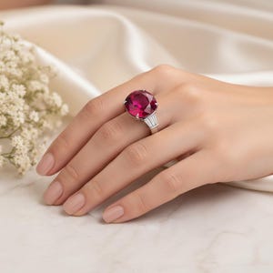 May include: A silver ring featuring a large, round, deep red gemstone. The ring has a classic design with three smaller, rectangular stones on each side of the main stone. The ring is displayed on a hand with manicured nails.
