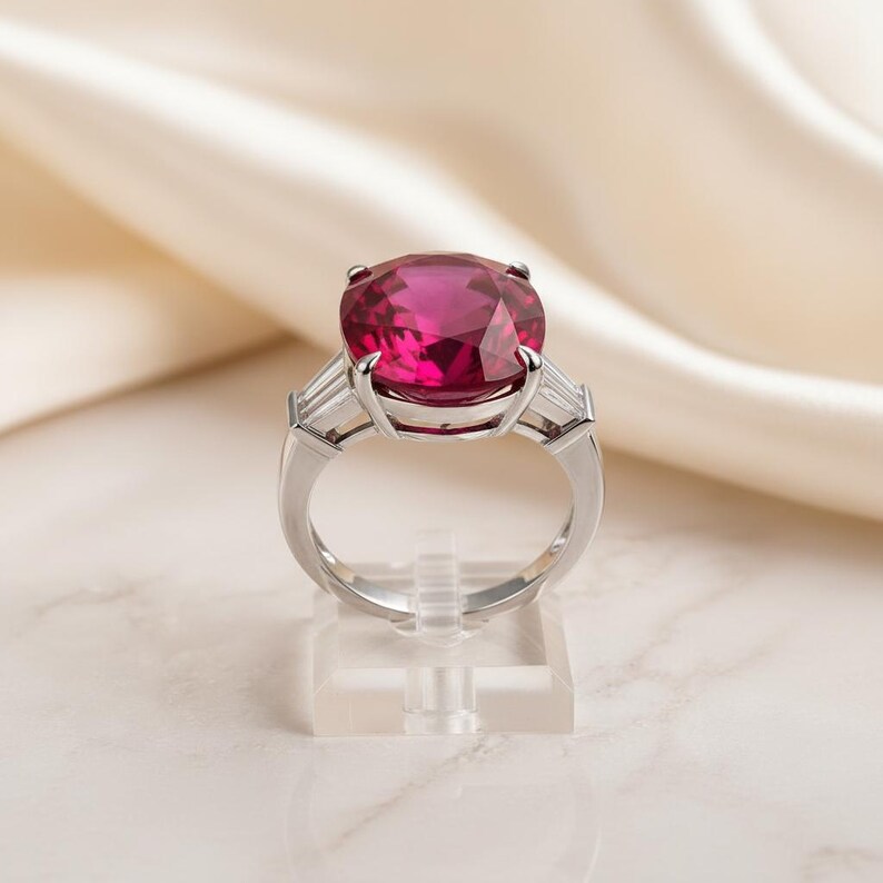Puede incluir: Un anillo de plata con una gran piedra preciosa rosa brillante de corte ovalado como pieza central. Dos piedras preciosas rectangulares y transparentes m&aacute;s peque&ntilde;as flanquean la piedra principal. El anillo se exhibe sobre un soporte transparente sobre un fondo blanquecino.