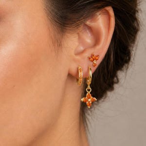 Può includere: Un set di orecchini color oro composto da un piccolo perno con tre pietre arancioni taglio marquise, un piccolo orecchino a cerchio con pietre arancioni pavé e un orecchino a cerchio con un ciondolo a fiore a quattro petali.