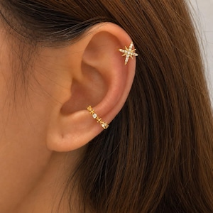 May include: A gold-toned ear cuff featuring a starburst design with embedded clear stones positioned on the upper cartilage, paired with a matching gold-toned ear cuff adorned with a series of small clear stones along the lower concha area.
