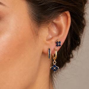 May include: A set of gold-toned earrings featuring a small stud with four blue stones arranged in a flower shape, a thin hoop earring with blue stones along the front, and a gold hoop earring with a dangling four-petaled blue stone charm.