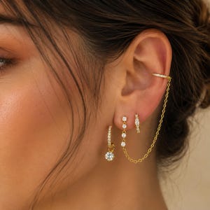 Könnte beinhalten: Ein Set aus goldfarbenen Ohrringen mit einer kleinen Ohrmanschette, die über eine feine Kette mit einem vertikalen Hänger mit drei klaren Steinen verbunden ist, sowie einem kleinen Creolen-Ohrring mit einem hängenden Stein und einer kleinen Pavé-Creole.