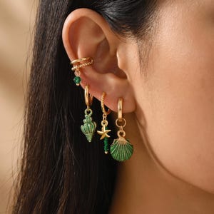 Puede incluir: Un conjunto de pendientes y un manguito de oreja dorados con dijes náuticos. La colección incluye una pequeña cuenta verde, una concha texturizada, una estrella de mar con cuentas verdes y un dije de concha en forma de abanico verde y dorado.