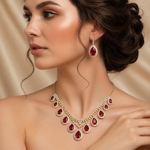 Puede incluir: Un conjunto de collar y pendientes dorados. Las joyas presentan piedras preciosas rojas en forma de lágrima, rodeadas de pequeños cristales transparentes. El conjunto está diseñado para un look elegante.