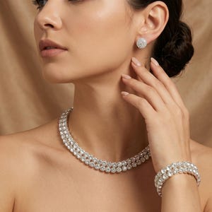 以下が含まれることがあります： ネックレス、ブレスレット、スタッドイヤリングが特徴の、まばゆいジュエリーセット。ネックレスとブレスレットは、複数の列の輝くクリアストーンで構成されています。イヤリングは丸く、お揃いの石のクラスターが付いています。ジュエリーはニュートラルな背景にセットされています。