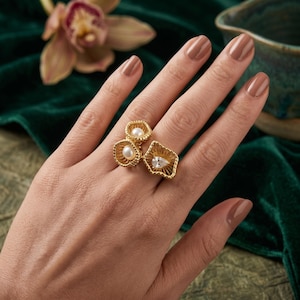 May include: A gold-toned ring featuring three distinct textured, scalloped-edge settings. Two of the settings contain small, round white pearls, while the third, larger setting holds a single, clear, pear-shaped faceted gemstone.