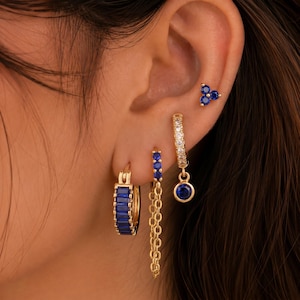 Könnte beinhalten: Ein Set aus goldfarbenen Ohrringen, bestehend aus einem kleinen Stecker mit drei blauen Steinen, einem Creolen-Ohrring mit blauem Anhänger, einem Creolen-Ohrring mit Kette und einem breiteren Creolen-Ohrring mit rechteckigen blauen Steinen.