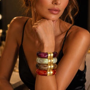 May include: A stack of four circular bangles featuring a marbled resin design in deep purple, pale green, brown, and orange. Each bangle is accented with a smooth, polished gold-tone metal rectangular connector piece that joins the two ends.