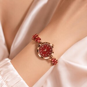 Puede incluir: Un reloj de cuarzo en tono oro rosa con esfera redonda roja y números arábigos blancos. La correa está compuesta por eslabones de metal en tono oro rosa, cada uno adornado con cristales rojos de talla marquesa en forma de hoja.