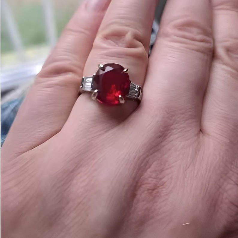Puede incluir: Un anillo plateado con una gran gema roja oscura de talla ovalada como pieza central. Dos gemas claras rectangulares m&aacute;s peque&ntilde;as flanquean la piedra principal. El anillo se muestra en una mano, mostrando el dise&ntilde;o y el color de la joya.