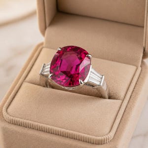 Puede incluir: Un anillo de platino con una gran piedra preciosa de color rojo rub&iacute; de talla ovalada como pieza central. El anillo est&aacute; flanqueado por dos piedras preciosas m&aacute;s peque&ntilde;as, rectangulares y transparentes. El anillo se muestra en un joyero beige.