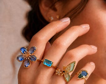 Conjunto de anillos ajustables en tono dorado, con flor de cristal azul, mariposa y gema aguamarina.