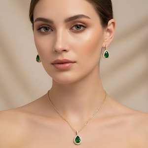 Può includere: Un set di gioielli color oro con collana e orecchini. Ogni pezzo ha una gemma verde smeraldo a forma di goccia, circondata da piccole pietre trasparenti. Il ciondolo della collana pende da una delicata catena e gli orecchini pendono dai lobi.