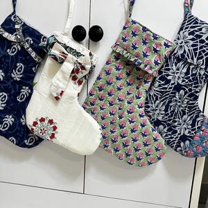 Puede incluir: Cuatro calcetines navideños acolchados colgados en fila. El primero es azul oscuro con patrones florales blancos. El segundo es blanco con un puño y punta de flores rojas y azules. El tercero es verde claro con flores rosas. El cuarto es azul oscuro con estampados florales blancos.