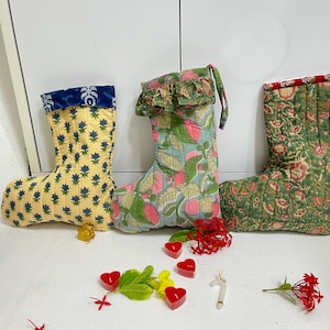 May include: Three quilted fabric stockings featuring floral and botanical patterns in yellow, light blue, and green. The stockings are decorated with small red heart-shaped candles, red flower clusters, a small white candy cane ornament, and a red bauble.