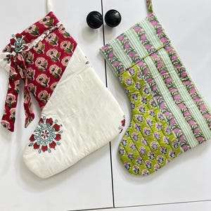 Op de afbeelding: Twee gewatteerde stoffen kerstsokken met ophanglussen. De linker sok heeft een rood bloemenpatroon op de boord en een witte onderkant met een centraal bloemmedaillon. De rechter sok heeft afwisselende groene gestreepte en bloemrijke panelen.