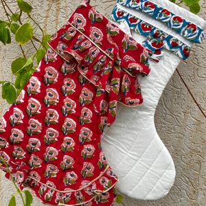 Pode incluir: Duas meias de Natal acolchoadas com alças para pendurar. Uma meia é vermelha com um padrão floral repetitivo em rosa e verde e acabamento com babados. A segunda meia é branca com um punho com babados em padrão floral azul, vermelho e verde.