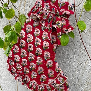 Puede incluir: Un calcetín navideño acolchado de tela roja intensa con un patrón floral repetitivo en tonos rosa, blanco y verde oscuro. El calcetín incluye adornos de volantes en el puño y la punta, rematados con un fino ribete de color claro.