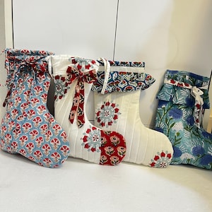 Puede incluir: Cuatro calcetines de tela acolchada con diversos estampados florales y de tipo mandala en tonos azul, rojo y blanco. Cada calcetín incluye un puño con volantes y una cinta de tela decorativa unida a la parte superior.