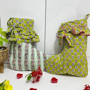 Könnte beinhalten: Zwei gesteppte Stoffstrümpfe mit einer lindgrünen Basis und einem wiederkehrenden rosa-weißen Blumenmuster. Der linke Strumpf hat vertikale grüne und weiße Streifen, beide verfügen über einen gerüschten Umschlag mit einer dekorativen Stoffschleife.