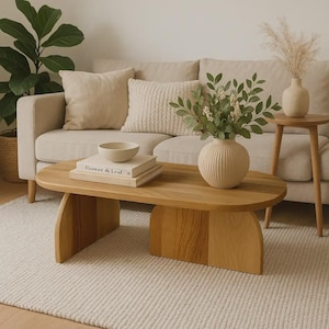 May include: A light oak oval coffee table with a distinctive base. The table is the focal point, set on a cream rug. A beige sofa, cushions, and decorative items, including a vase and books, are in the background. The book reads "Flower & Leaf."