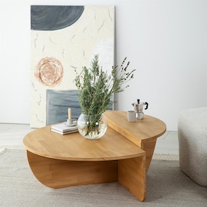Op de afbeelding: Een licht houten salontafel met een uniek, tweedelig ontwerp. Een glazen kom met groen staat op de tafel, samen met een kleine kaars, een boek en een koffiezetapparaat. De tafel staat voor een groot abstract schilderij.