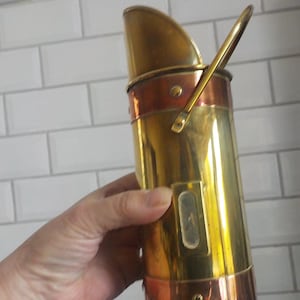 May include: A cylindrical brass and copper coal scuttle featuring a slanted top opening, a metal handle attached to the side, and a small rectangular glass viewing window on the front body. The vessel is constructed with polished brass panels and copper bands.