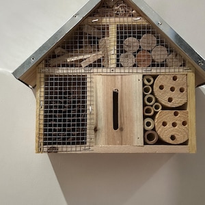 May include: A wooden insect hotel with a peaked metal roof, featuring multiple compartments filled with natural materials including pine cones, hollow bamboo reeds, wooden blocks with drilled holes, and wood shavings, all secured behind a wire mesh.