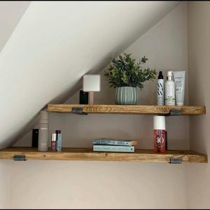 May include: Two wooden shelves with a rustic finish, fixed to a wall with black metal brackets. The top shelf displays a potted plant, beauty products, and a small mirror. The bottom shelf holds books and more beauty products.