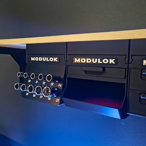 May include: Black MODULOK storage units mounted under a wooden shelf. The units have drawers and numbered tool holders. The text "MODULOK" is displayed on each unit. The tool holders have numbers from 10 to 21.