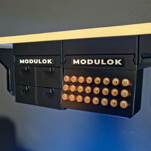May include: Black MODULOK battery organizer with four small drawers on the left and a section for holding AA batteries on the right. The organizer is mounted under a wooden shelf. The word MODULOK is printed in white.