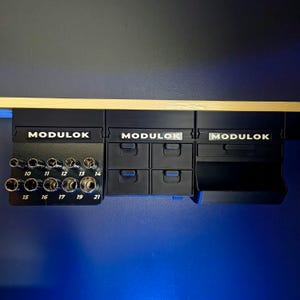 May include: A black Modulok wall-mounted storage system featuring three modular units. The left unit holds ten metal sockets labeled 10 through 21. The center unit contains four small drawers, and the right unit features an open storage bin.