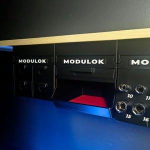 以下が含まれることがあります： 白字で「MODULOK」と書かれた黒いモジュール式収納システム。引き出しとコンパートメントがあり、開いた部分には赤い内装が見えます。数字の10、11、15、16が表示されています。