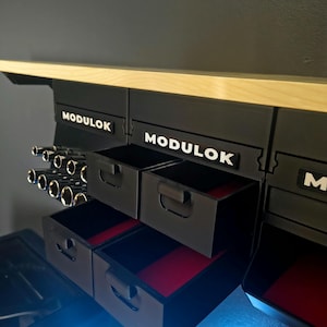 May include: Black modular storage unit with drawers and tool holders. The unit has the word "MODULOK" in white lettering. The drawers have red felt lining. The unit is mounted under a light-colored wooden shelf.