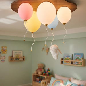 May include: A whimsical ceiling light fixture shaped like a cloud, with five balloon-shaped lights in pink, yellow, white, and blue. The fixture is made of wood and hangs in a child's bedroom.
